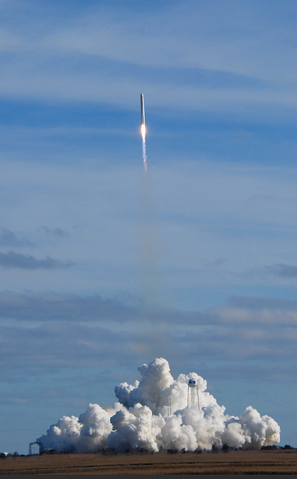 A launched rocket soars above billowing white clouds of exhaust, white-hot flame shooting from its stern, against a partly cloudy blue sky.