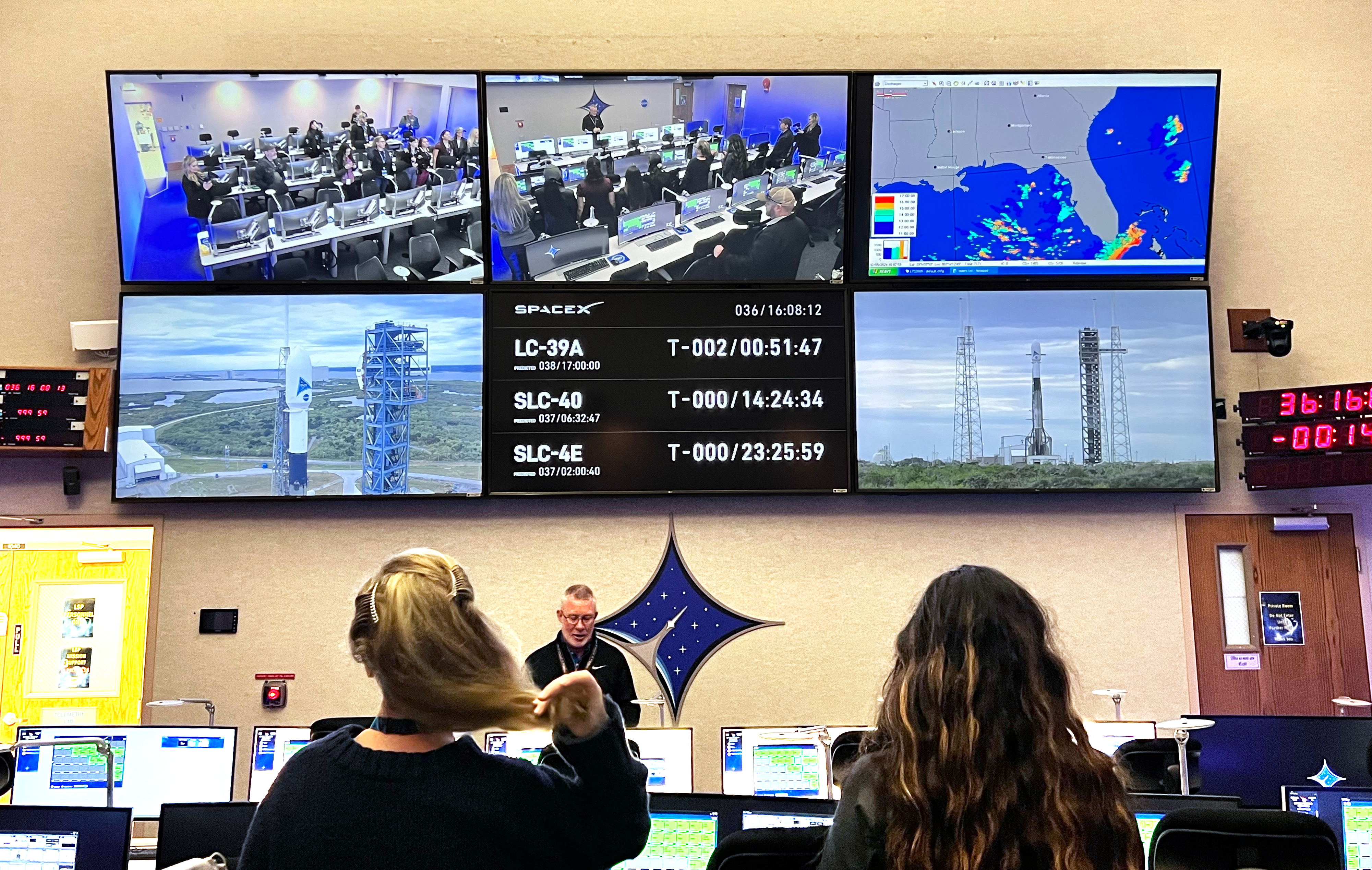 Two women facing away from the camera in a large mission-control room. Before them are a man in glasses and six video screens showing camera views of a rocket on a launch pad, weather maps, and other mission data.